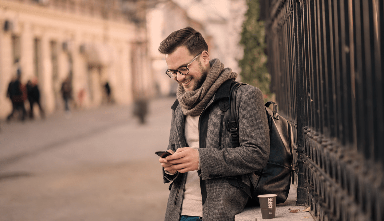 A man standing beside the wall drinking coffee and smiling while looking at his mobile