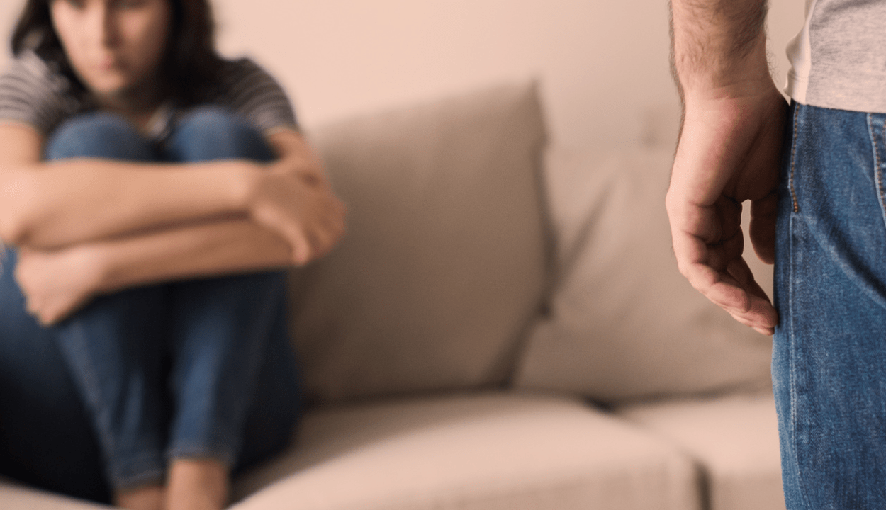 A woman sitting on a sofa looking upset and a man standing in front of him